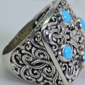 May include: A silver ring with an ornate, swirling design and five round, light blue stones. The ring has a wide band and a detailed, floral-inspired pattern. The stones are evenly spaced across the top of the ring.
