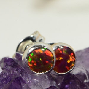 May include: A pair of silver stud earrings featuring round, iridescent opal gemstones. The opals display a range of red, orange, green, and purple colours.