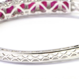 May include: A silver bangle bracelet with a delicate, intricate design. The bracelet is set with a row of red gemstones.