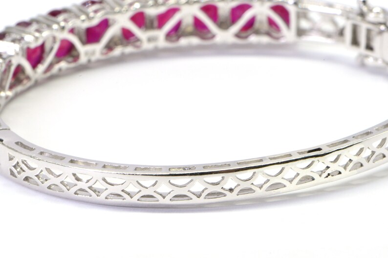 May include: A silver bangle bracelet with a delicate, intricate design. The bracelet is set with a row of red gemstones.