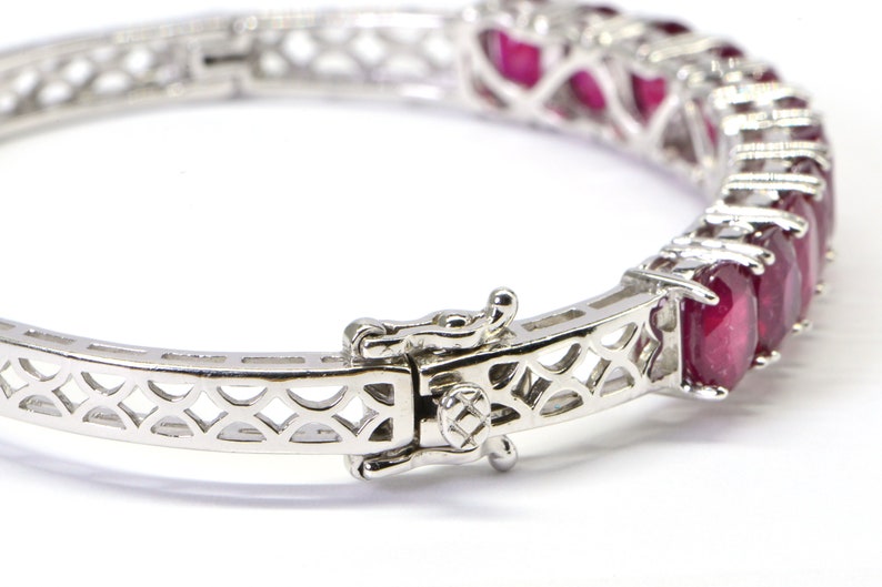 May include: A silver bangle bracelet with a delicate, openwork design and a row of red ruby gemstones.