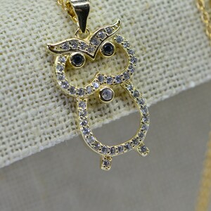 Puede incluir: Un collar de oro con un colgante de búho con diamantes de imitación transparentes y negros. El búho está rodeado de diamantes de imitación transparentes y tiene diamantes de imitación negros para los ojos.