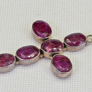 May include: A silver cross pendant with five oval-shaped ruby gemstones. The pendant has a loop at the top for attaching to a chain.