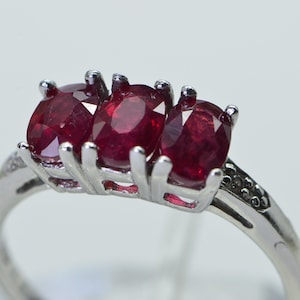 May include: A silver ring with three red ruby stones set in a prong setting. The ring has a band of small diamonds.