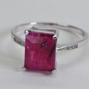 May include: A silver ring featuring a large, rectangular, pink ruby gemstone set in a prong setting. The band is adorned with small, clear gemstones.
