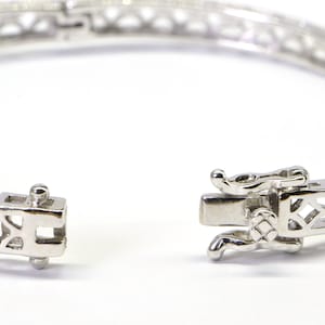 May include: A silver bangle bracelet with a delicate openwork design and a hinged clasp. The bracelet is adorned with small, red gemstones.