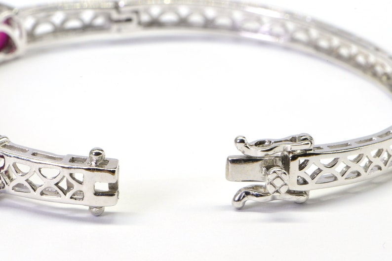 May include: A silver bangle bracelet with a delicate openwork design and a hinged clasp. The bracelet is adorned with small, red gemstones.