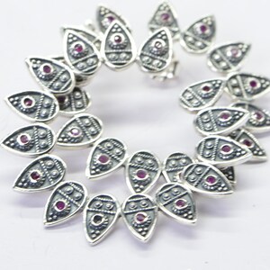 May include: A silver charm bracelet with a teardrop design. Each teardrop charm has a small pink gemstone in the center and intricate detailing around the edges.