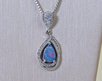 May include: A silver-coloured pendant necklace featuring a pear-shaped dark blue opal with iridescent flashes of red and green. The opal is surrounded by a swirling frame of small clear stones, with a matching stone-encrusted bail on a box chain.