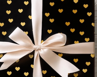 May include: A wrapped gift with black paper covered in gold hearts. A cream-colored ribbon is tied in a bow around the package. The gift is on a white wooden surface.