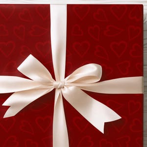 Könnte beinhalten: Eine verpackte Geschenkbox mit rotem Hintergrund und einem Muster aus Herzen. Die Box ist mit einem cremefarbenen Band und einer Schleife gebunden. Die Geschenkbox befindet sich auf einer weißen Holzoberfläche.