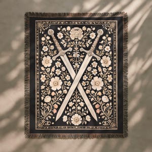 Pode incluir: Cobertor preto com um design floral e duas espadas cruzadas. O cobertor apresenta um padrão floral creme sobre um fundo preto, com uma borda com franjas. As espadas são prateadas.