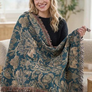May include: A woven throw blanket featuring a deep teal background with a prominent gold and tan floral damask pattern. The rectangular textile has a textured appearance with decorative fringe detailing along the edges, showcasing a dense weave.