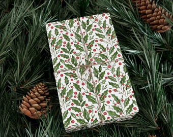 May include: A wrapped gift with a festive holly and berry pattern, tied with twine, rests on a bed of evergreen branches and pine cones. The wrapping paper features a white background with green leaves and red berries, perfect for the holiday season.