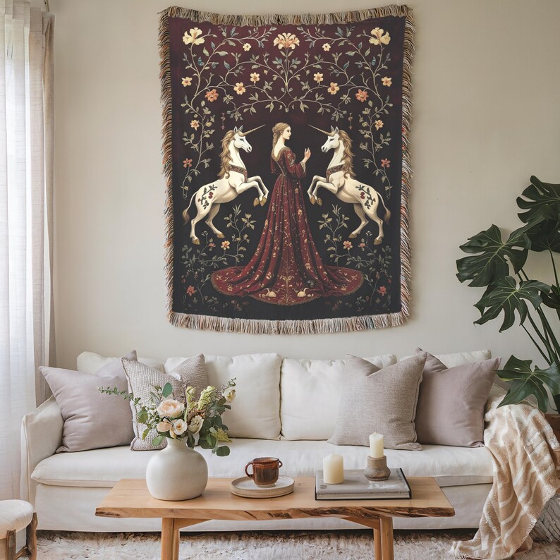 Puede incluir: Un tapiz que representa a una mujer con un vestido rojo entre dos unicornios blancos. El tapiz tiene un fondo rojo oscuro con detalles florales y de vid. Cuelga sobre un sof&aacute; blanco con cojines y una mesa de centro de madera.