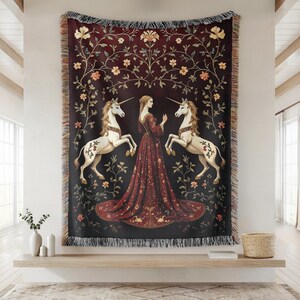 Puede incluir: Una manta de tapiz que representa una escena medieval. Muestra a una mujer con un vestido rojo entre dos unicornios blancos. El fondo es rojo oscuro con motivos florales y los bordes tienen un borde con flecos.