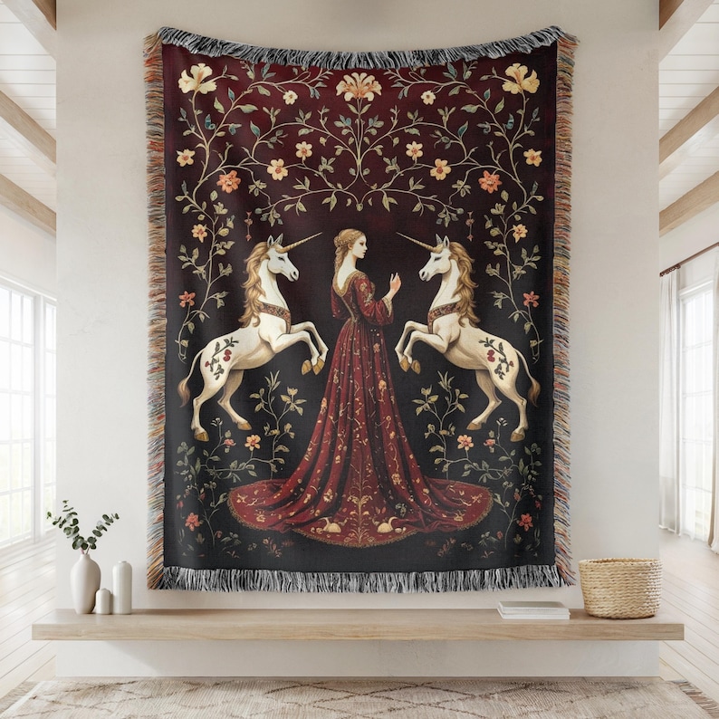 Puede incluir: Una manta de tapiz que representa una escena medieval. Muestra a una mujer con un vestido rojo entre dos unicornios blancos. El fondo es rojo oscuro con motivos florales y los bordes tienen un borde con flecos.