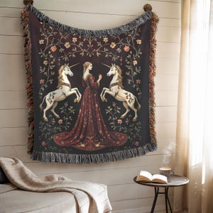 Puede incluir: Una manta de tapiz que representa una escena medieval con una mujer con un vestido rojo, dos unicornios blancos y detalles florales sobre un fondo oscuro. La manta tiene un borde con flecos y est&aacute; colgada en una pared.