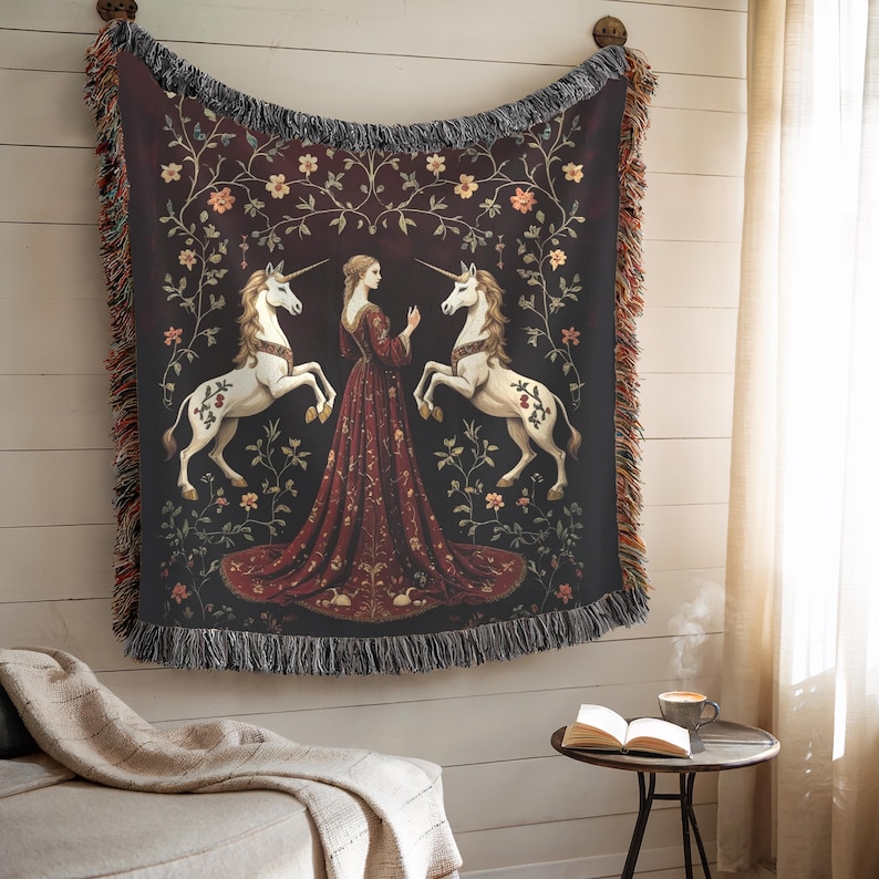 Puede incluir: Una manta de tapiz que representa una escena medieval con una mujer con un vestido rojo, dos unicornios blancos y detalles florales sobre un fondo oscuro. La manta tiene un borde con flecos y est&aacute; colgada en una pared.