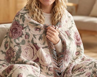 May include: A woven throw blanket featuring a repeating floral pattern with muted rose-pink flowers and sage green foliage on a cream-colored background, finished with a multi-colored fringed edge along the side of the fabric.