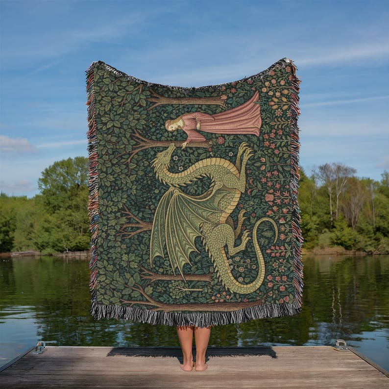 May include: A woven tapestry blanket featuring a green dragon with outstretched wings and a woman in a pink dress. The blanket has a dark green background with a floral and tree design. The blanket has a fringed edge.
