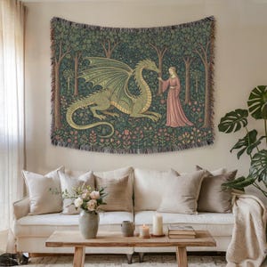 May include: A woven tapestry featuring a green dragon and a woman in a pink dress in a forest scene. The dragon is lying down with its wings spread. The tapestry is displayed above a white sofa with beige cushions.