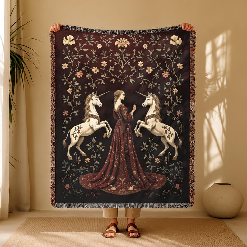 Puede incluir: Una manta tejida con una mujer y dos unicornios. El dise&ntilde;o est&aacute; sobre un fondo burdeos oscuro con detalles florales y de vid. La manta tiene un borde con flecos.
