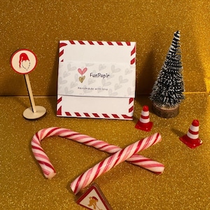Könnte beinhalten: Eine festliche Anordnung mit Zuckerstangen, Miniatur-Leitkegeln, einem kleinen Weihnachtsbaum und einem Schild mit einer Weihnachtsmann-Illustration. Eine weiß-rote Box mit dem Text "FunPapir" ist ebenfalls vorhanden. Der Hintergrund ist eine glitzernde goldene Oberfläche.