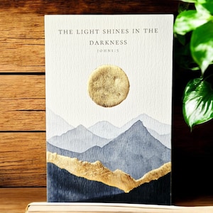 May include: A watercolour art print depicting a mountain landscape with a gold sun and accents. The text "THE LIGHT SHINES IN THE DARKNESS JOHN 1:5" is printed at the top. The artwork has a textured paper appearance.