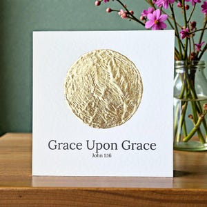 Könnte beinhalten: Eine weiße Karte mit dem Text "Grace Upon Grace" und "John 1:16" in Schwarz. Ein strukturierter goldener Kreis befindet sich über dem Text. Rosa Blumen in einer Glasvase sind im Hintergrund.