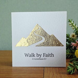 Könnte beinhalten: Eine weiße Grußkarte mit einem goldfolierten Bergdesign und einem gewundenen Pfad. Der Text "Walk by Faith" und die Referenz "2 Corinthians 5:7" sind unter der Bergillustration in Schwarz gedruckt.