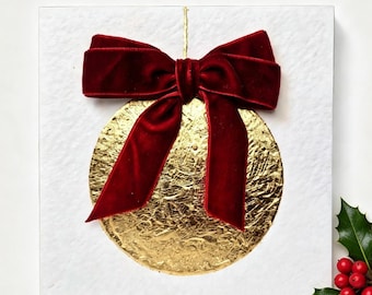 May include: A square white textured card featuring a circular gold foil ornament design centered in the middle. A deep red velvet ribbon is tied in a bow at the top of the gold circle. A small sprig of green holly leaves with red berries is visible.