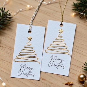 May include: Two rectangular white gift tags featuring a gold foil-stamped continuous line drawing of a Christmas tree topped with a star. Below the tree, the text Merry Christmas is printed in black script. One tag has a white and gold cord.