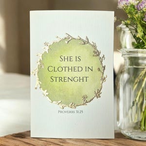 Op de afbeelding: Een wenskaart met de tekst "SHE IS CLOTHED IN STRENGHT" in een gouden bloemenkrans. De achtergrond is een verloop van groen en wit. De onderkant van de kaart luidt "PROVERBS 31:25".