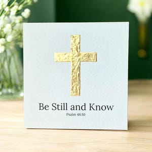 Könnte beinhalten: Eine weiße Karte mit einem goldenen Kreuz in der Mitte. Unter dem Kreuz steht der Text "Be Still and Know", darunter "Psalm 46:10". Die Karte liegt auf einer Holzoberfläche.