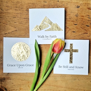 May include: Three white cards with gold foil designs and text. One card features a mountain with a path, the text "Walk by Faith". Another has a gold circle with "Grace Upon Grace". The last card has a gold cross with "Be Still and Know". A tulip is in the foreground.