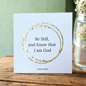 Peut inclure: Une carte carrée bleu clair avec du texte doré et un motif circulaire doré. Le texte dit "Be Still, and Know that I am God" avec "Psalm 46:10" en dessous. La carte est posée sur une surface en bois.