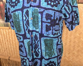 Op de afbeelding: Blauw overhemd met korte mouwen met een herhalend geometrisch patroon in blauw- en tinten. Het patroon bevat gestileerde tiki-figuren en bloemvormen. Het overhemd heeft een klassieke kraag en houten knopen.