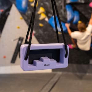 May include: A light purple rectangular climbing hangboard featuring a central 25mm edge, suspended by a black cord. The training tool has a smooth, matte finish and a recessed grip area designed for finger strength training in a climbing gym.