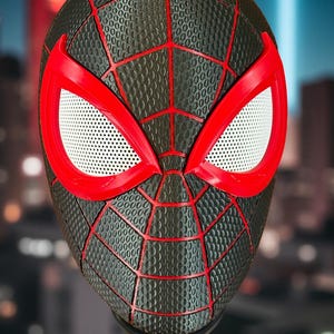 May include: A black and red Spider-Man mask with a textured surface and a web design. The mask features red eye frames with white mesh eye coverings. The mask is displayed on a black stand, with a blurred cityscape in the background.
