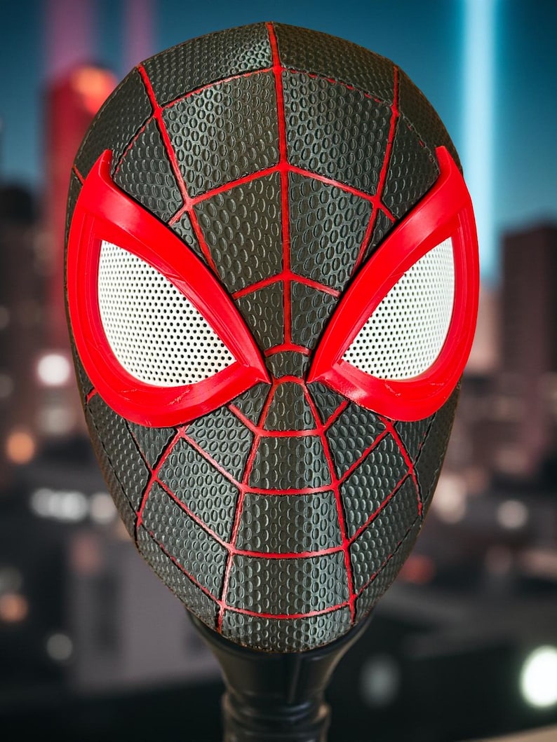 May include: A black and red Spider-Man mask with a textured surface and a web design. The mask features red eye frames with white mesh eye coverings. The mask is displayed on a black stand, with a blurred cityscape in the background.