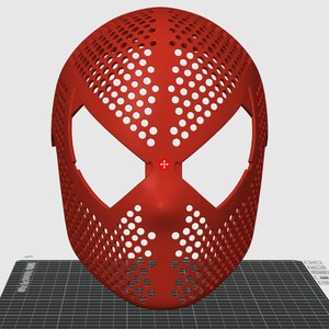 May include: A red, perforated mask with large eye openings and a distinctive design. The mask is likely for cosplay or costume use, and is made of a solid material. The mask is displayed on a black and grey surface.