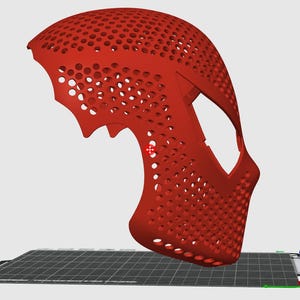 May include: A red, 3D-printed mask with a honeycomb pattern. The mask has a curved design with eye openings and a chin piece. It is resting on a black and grey surface.