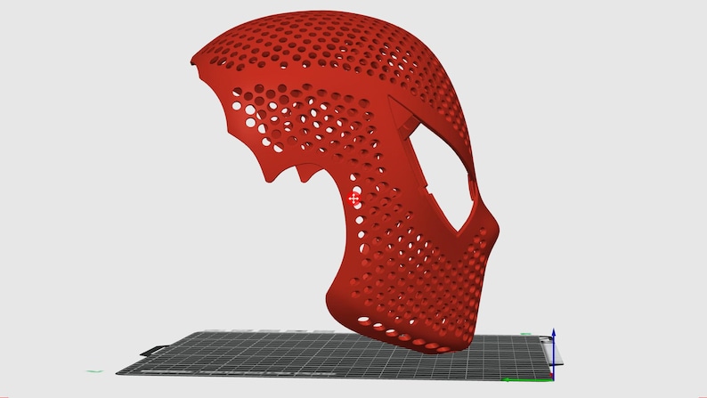 May include: A red, 3D-printed mask with a honeycomb pattern. The mask has a curved design with eye openings and a chin piece. It is resting on a black and grey surface.