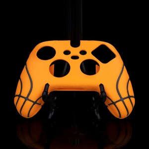 Puede incluir: Una funda de silicona naranja para mando con detalles negros de baloncesto. La funda tiene recortes para botones y joysticks, diseñada para adaptarse a un mando de juegos. El fondo es negro.