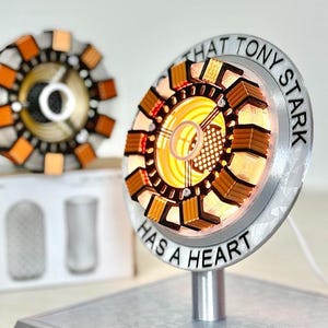 May include: A replica Arc Reactor, a circular device with a silver rim and a yellow and orange core. The text "THAT TONY STARK HAS A HEART" is printed on the rim. Another reactor is visible in the background. The device is on a silver stand.