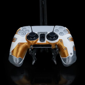May include: A custom video game controller in white, gold, and grey. The controller has gold accents on the grips and grey detailing. The buttons and joysticks are black. It is displayed on a black stand against a black background, highlighting its design.