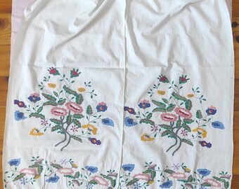 Puede incluir: Tela blanca con bordados florales en rosa, azul, amarillo y verde. El bordado presenta un diseño floral detallado. El borde inferior tiene un ribete de encaje blanco. La tela está sobre una superficie rosa.