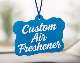 Può includere: Un deodorante per auto personalizzato blu a forma di nuvola, appeso allo specchietto retrovisore di un'auto. Le parole "Custom Air Freshener" sono scritte in bianco. Il cruscotto e lo specchietto retrovisore dell'auto sono visibili sullo sfondo.