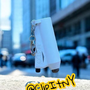 May include: A white, rectangular Clip It device with a keychain attachment. The device has a black base and the text "CLIP IT" is visible. The image also includes the text "@clipItNY" in a yellow circle.